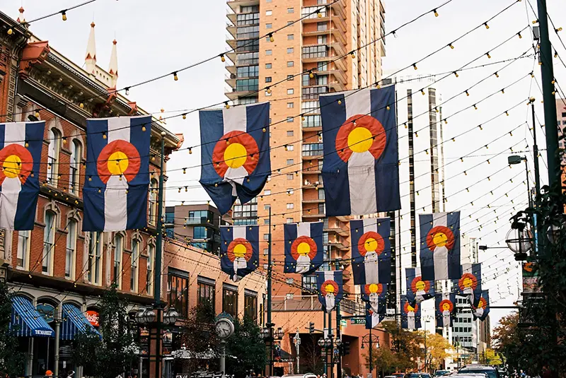Colorado state flags hang above a city street lined with historic buildings, restaurants, and shops, with string lights overhead and tall modern buildings in the background on a cloudy day.