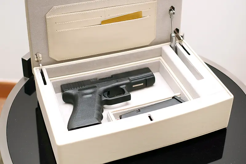 A black handgun is securely placed inside a beige lockbox, which is open on a black table. The lockbox also contains a magazine and some compartments, including a slot with a gold card.