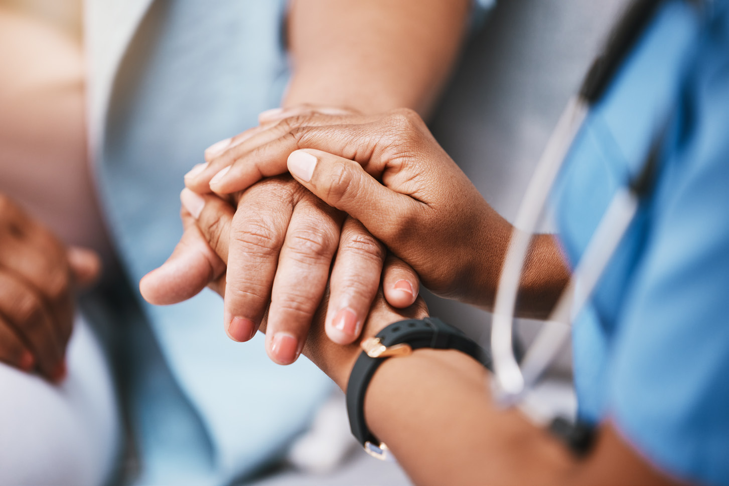 A healthcare worker gently holds a patients hand, conveying comfort and support. The patient’s hands rest on top, while the worker wears blue scrubs and a stethoscope. Only their hands and arms are visible.