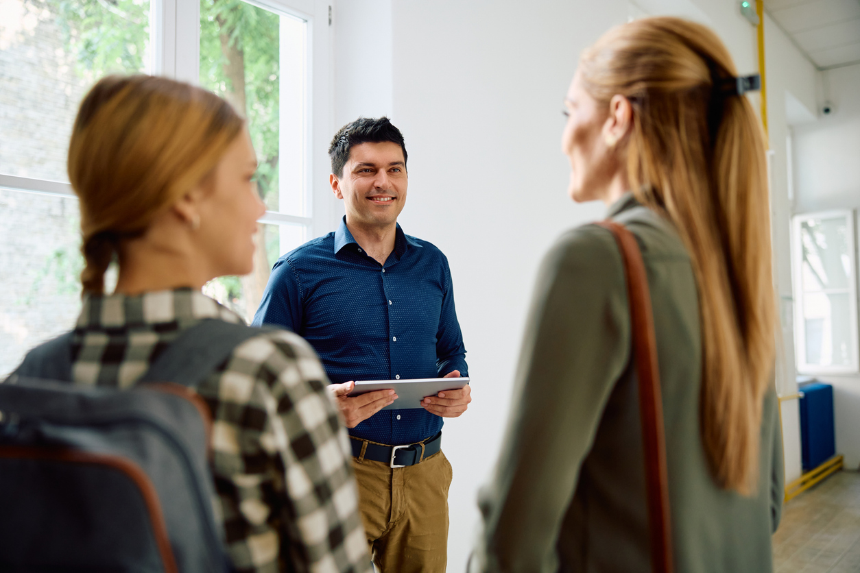 A man holding a tablet smiles and talks to two women, one with a backpack, in a brightly lit room with large windows and white walls.