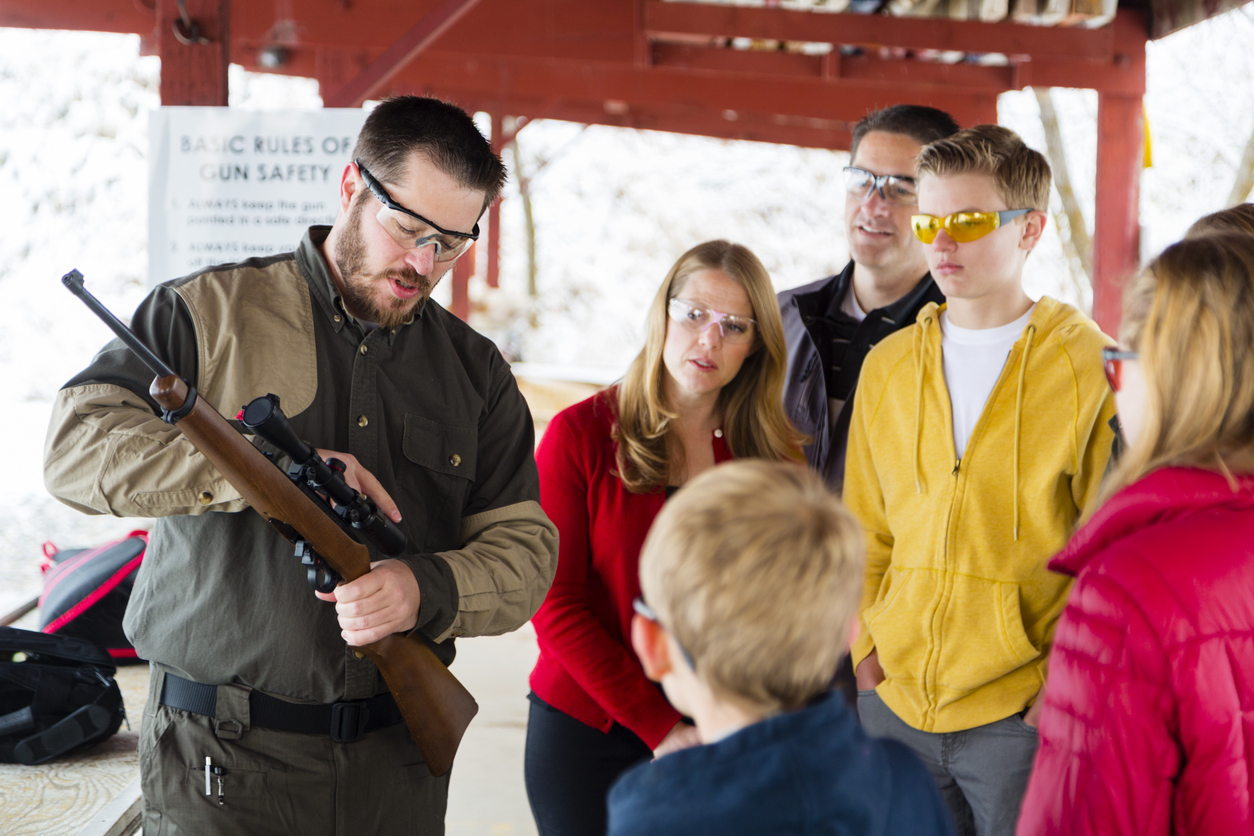 A man demonstrates how to use a rifle to a group of adults and children wearing safety glasses at an outdoor shooting range. A gun safety sign is visible in the background.