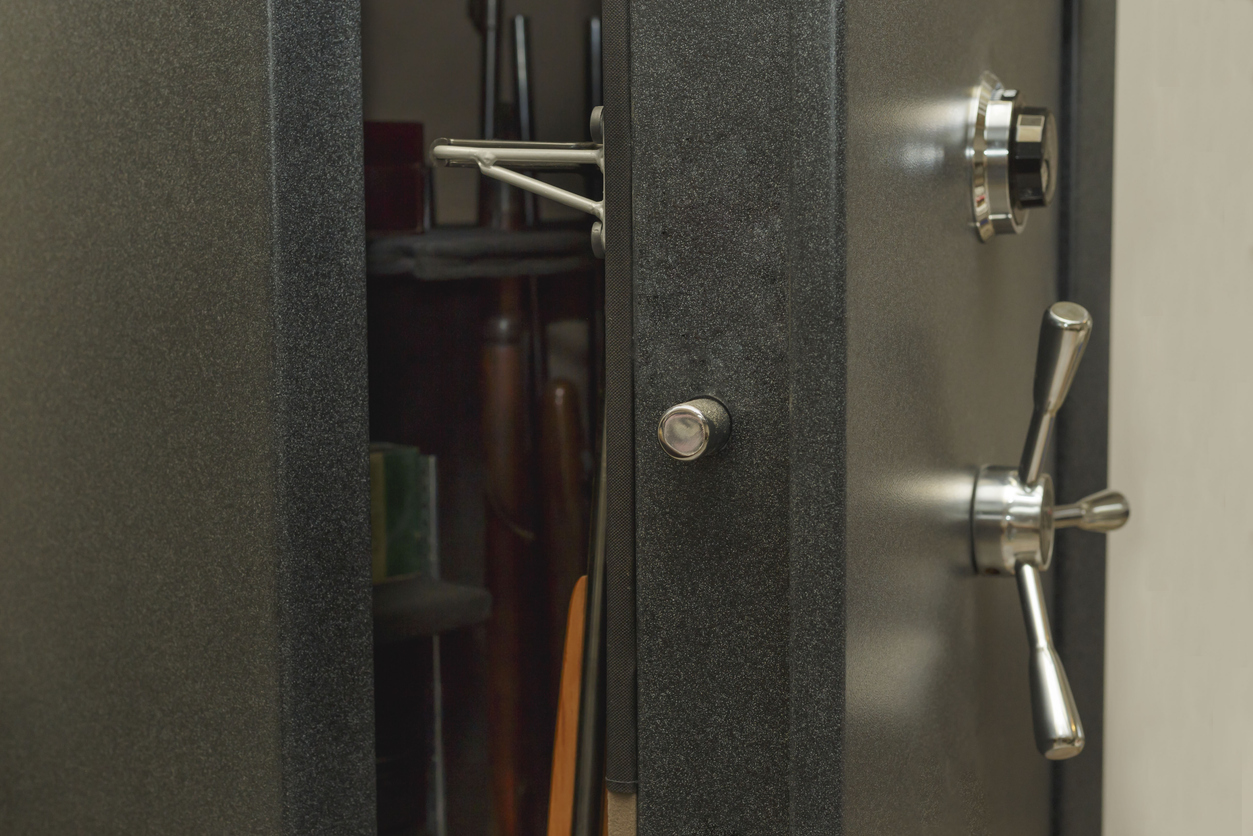 A partially open gray safe reveals several firearms and objects stored inside. The safe door features a combination lock and chrome handle levers.