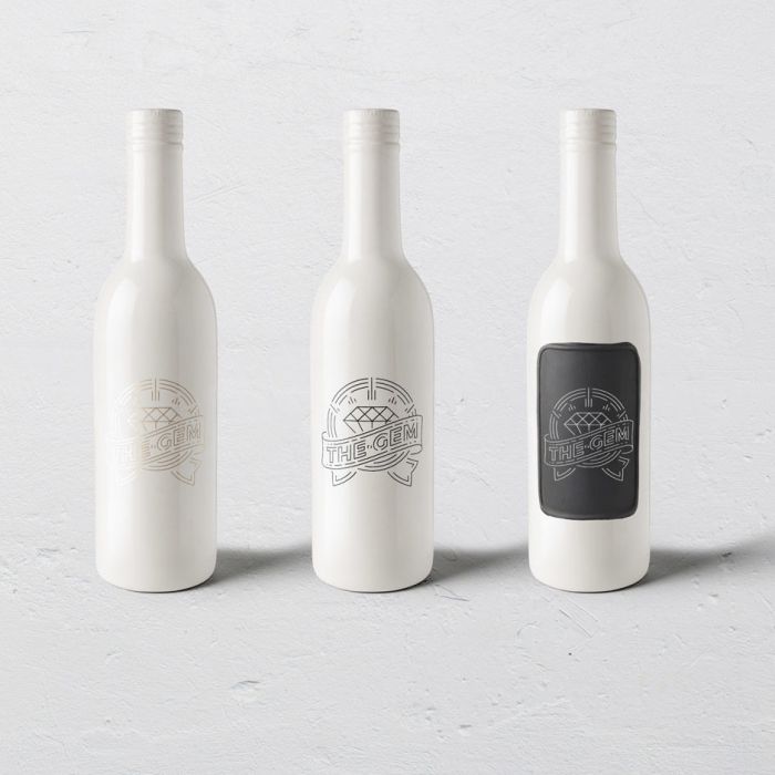 Three white wine bottles with cork tops are displayed side by side against a light gray background. Each bottle features a logo; the middle bottle has a clearer logo, and the right bottle has a black label with white design.