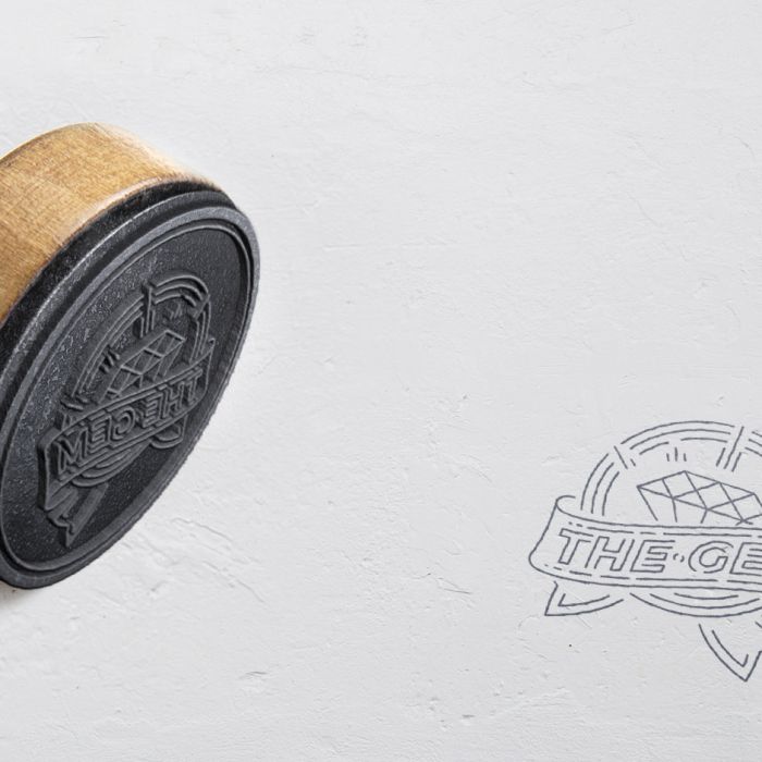 A rubber stamp with a wooden handle lies on its side, next to its stamped imprint on white paper. The stamp design features a diamond and the text “THE GEM” inside a ribbon banner.