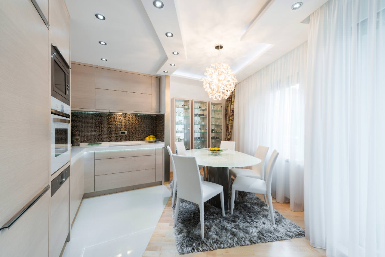 Modern kitchen and dining area with light wood cabinets, white appliances, round table with five white chairs on a gray rug, glass display cabinet, and a decorative chandelier, brightened by natural light from large windows.