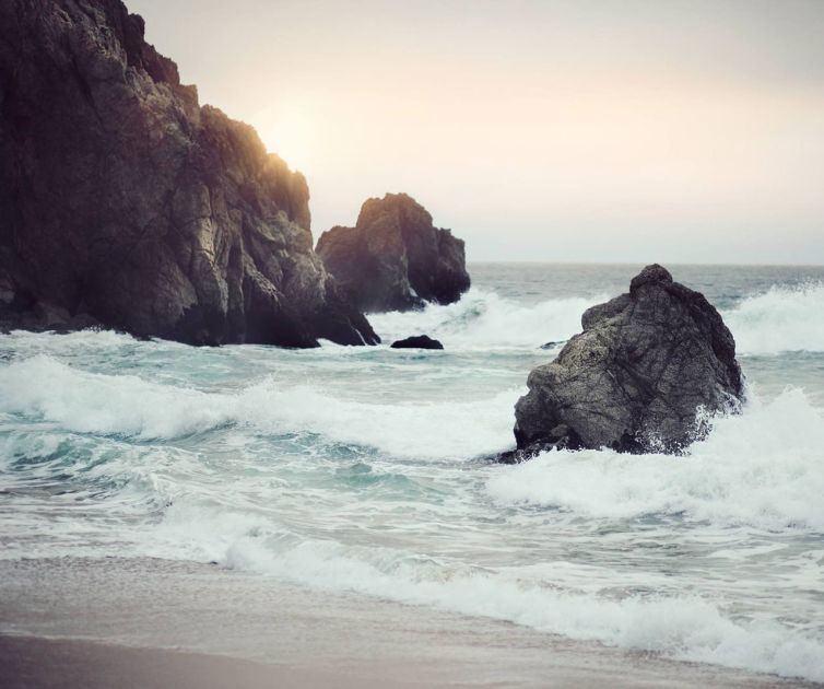 Waves crash against large rocky formations along a sandy shoreline at sunset, with soft sunlight partially obscured by clouds and rocks in the background.