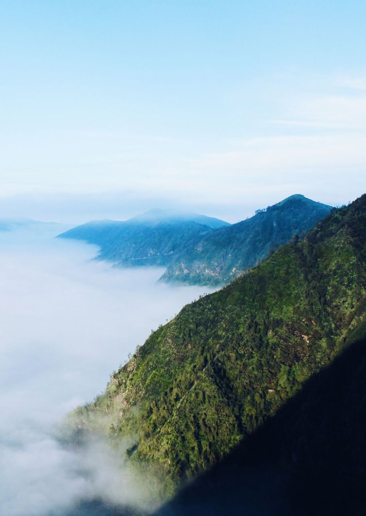 Green mountains partially covered with mist and clouds, under a clear blue sky, creating a serene and tranquil landscape.