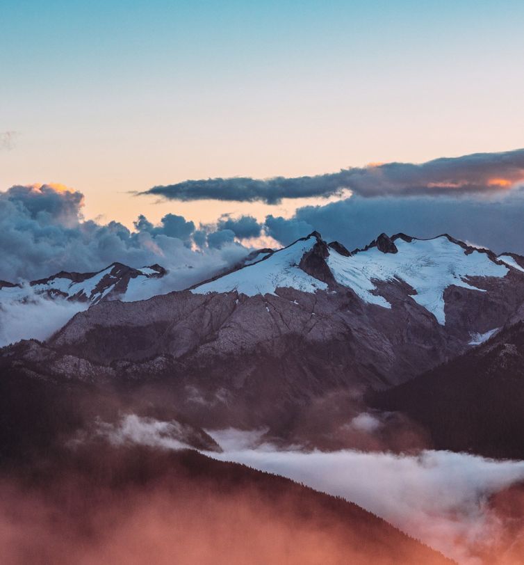 Snow-capped mountains under a colorful sky at sunrise or sunset, with clouds and mist drifting through the valleys and forested hills in the foreground.
