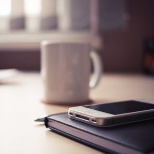 A smartphone rests on a closed notebook with a white coffee mug in the background on a light-colored table, softly lit by natural light.