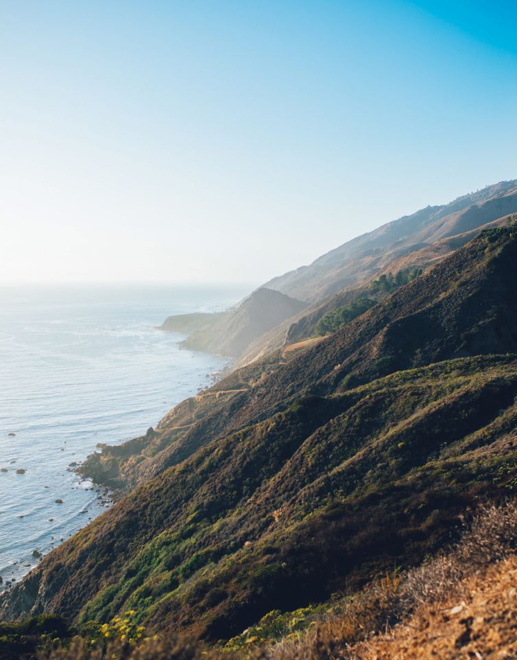Steep, rugged coastal cliffs covered in greenery slope down to the blue ocean under a clear sky, with gentle sunlight illuminating the scene.
