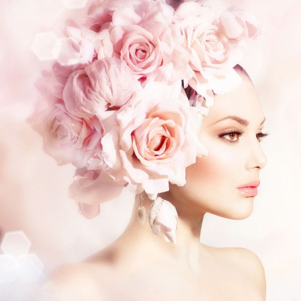 A woman with flawless makeup wears a large, elaborate headdress made of pale pink roses and peonies, set against a soft, dreamy pastel background. Her expression is calm and elegant.