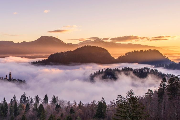 A scenic sunrise view of misty hills and forest, with mountains in the background and a church tower partially visible above the fog.