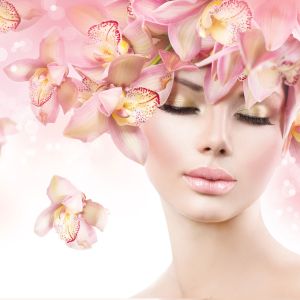 A woman with closed eyes and soft makeup is adorned with pink orchids in her hair against a light, dreamy background, creating an ethereal and floral beauty theme.