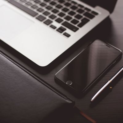 A close-up of a laptop keyboard, a smartphone, a closed notebook, and a pen arranged neatly on a dark desk.