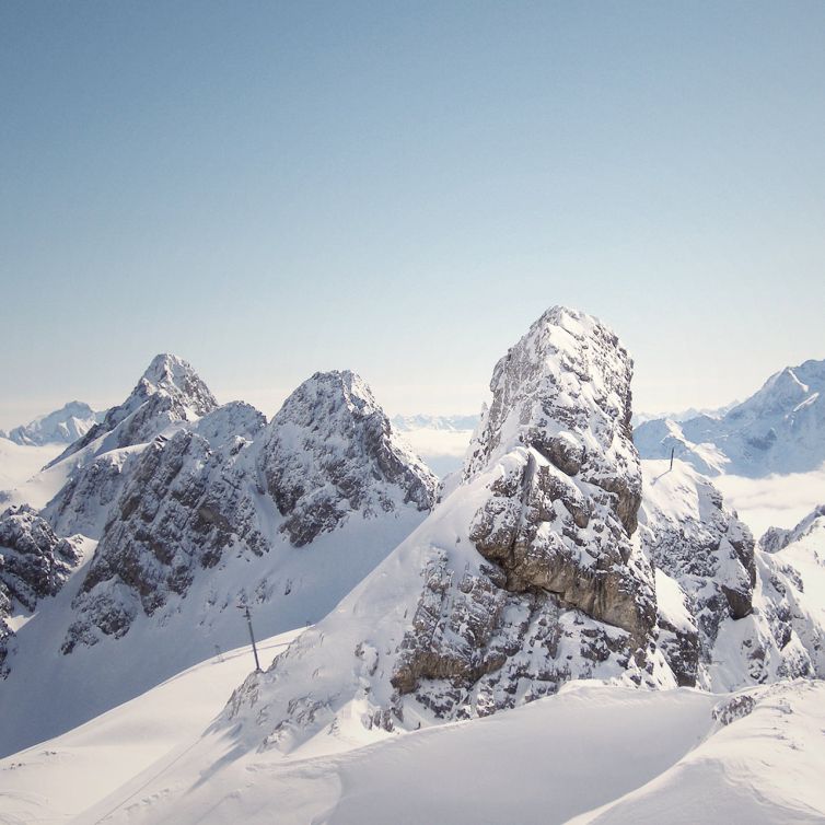 Snow-covered mountain peaks under a clear blue sky, with sunlight highlighting the rocky surfaces and gentle slopes. The distant mountains fade into the horizon, creating a serene and peaceful winter landscape.