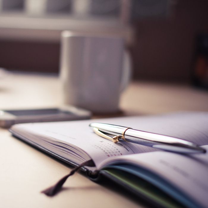A close-up of an open notebook with a pen resting on its pages, a blurred mug, and a smartphone in the background on a light-colored desk.