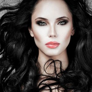 A woman with long, wavy black hair, striking blue eyes, bold eye makeup, and glossy red lips gazes directly at the camera against a neutral background.