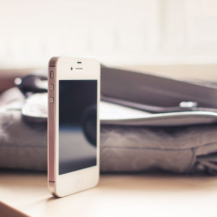 A white smartphone with a black screen stands upright on a light surface next to a gray bag, with soft natural light coming from the background.