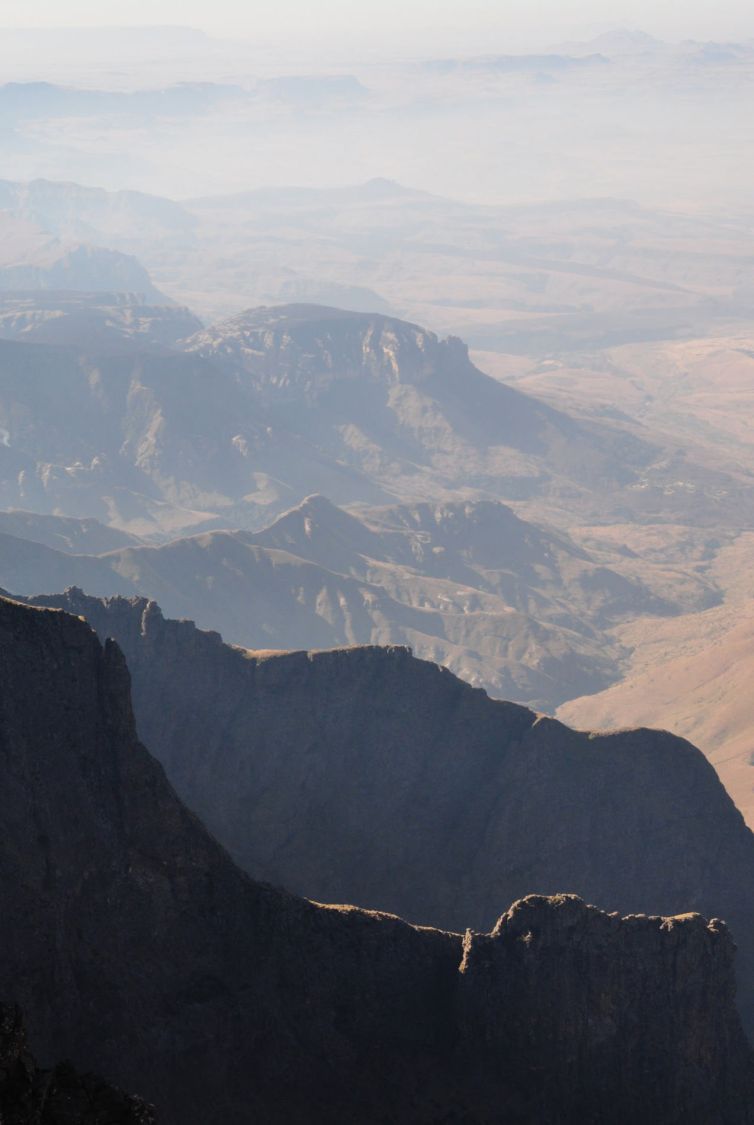 A dramatic, hazy landscape view of layered mountains and deep valleys, with sunlight casting shadows and illuminating the ridges in the foreground. The scene fades into the misty distance.