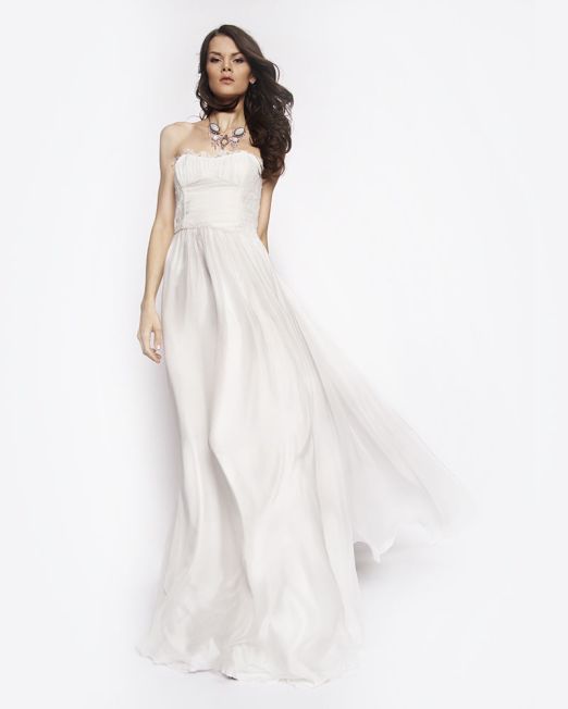 A woman wearing a strapless, flowing white gown poses against a plain white background. She has long, wavy hair and is accessorized with a statement necklace.