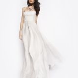 A woman wearing a strapless, flowing white gown poses against a plain white background. She has long, wavy hair and is accessorized with a statement necklace.