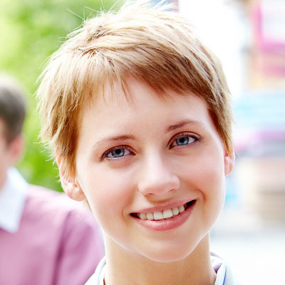 A young person with short, light brown hair smiles warmly at the camera. The background is blurred with hints of greenery and colorful shapes.