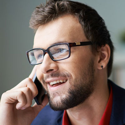 A smiling man with glasses and a beard is talking on a smartphone. He is wearing a dark jacket and a red shirt, and has a small earring in his left ear.