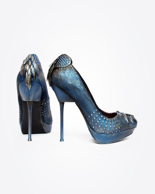 A pair of high-heeled shoes in metallic blue, decorated with small studs and featuring layered, scale-like accents on the heels. The shoes have a fantasy-inspired, ornate design.