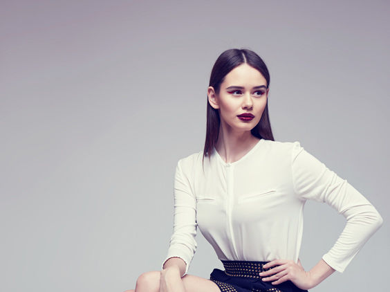 A young woman with straight brown hair, dark lipstick, and a white long-sleeve blouse sits with one hand on her hip, looking to the side against a neutral background.