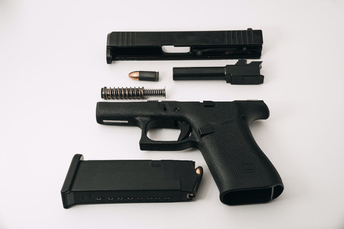 A disassembled black handgun is laid out on a white surface, showing the slide, barrel, recoil spring, magazine, and frame with a single bullet visible in both the barrel and the magazine.