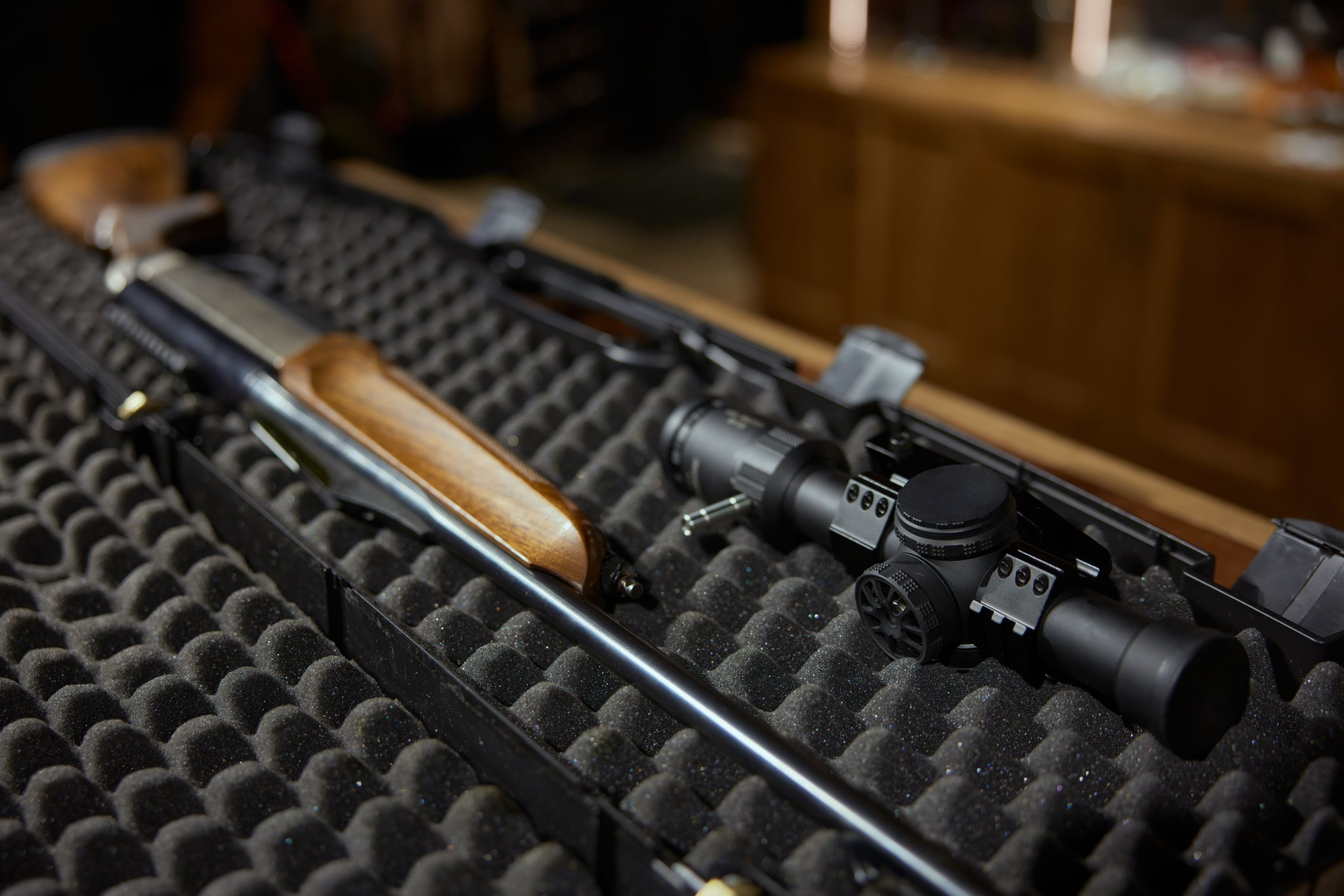 A rifle with a wooden stock and scope is placed inside a black foam-lined hard case; the background is out of focus, showing wooden furnishings.