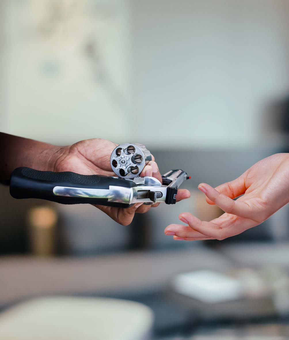 A person holding an open revolver hands it to another person, whose hand is reaching out to take it. The background is blurred.