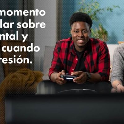 A young man and woman sit on a couch smiling and playing video games. Spanish text on the left reads, El mejor momento para hablar sobre salud mental y armas es cuando no hay presión. A Let’s Talk Guns Colorado logo is in the corner.