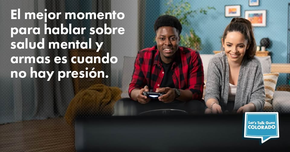 A young man and woman sit on a couch smiling and playing video games. Spanish text on the left reads, El mejor momento para hablar sobre salud mental y armas es cuando no hay presión. A Let’s Talk Guns Colorado logo is in the corner.